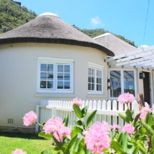 A small white house with a fence and pink flowers at Amber Light Wilderness Beach Cottage in Wilderness