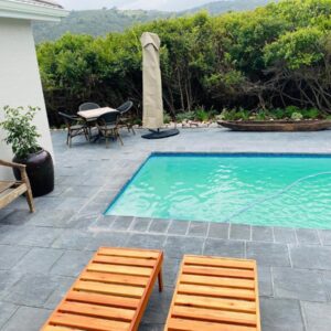 A swimming pool with two wooden chairs and a table at Mes Amis Beach Guest House in Wilderness