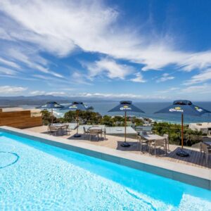 A swimming pool with chairs and umbrellas and the ocean at Plett Quarter Hotel - Lion Roars Hotels & Lodges in Plettenberg Bay