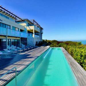 A swimming pool next to a building with chairs and the ocean at The Shark Bay Villas in Langebaan